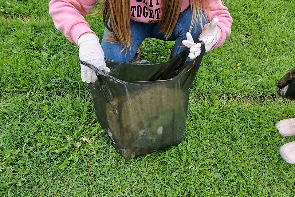 Ett barn med vita handskar står på huk på gräsyta med en halvfull plastpåse 