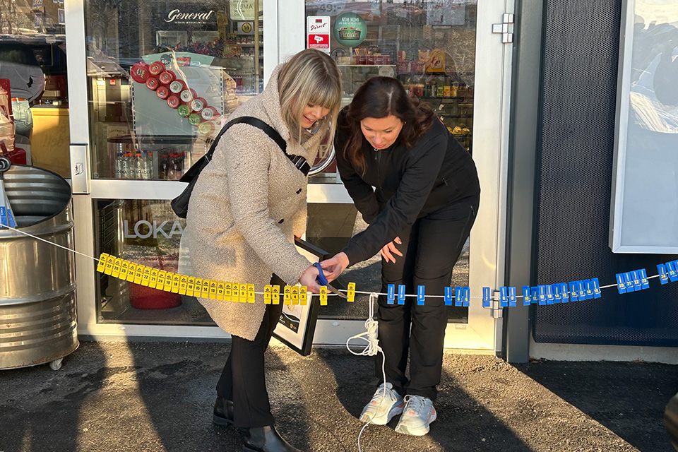 Två kvinnor håller tillsammans i en sax och klipper ett band med nypor för lotter. Utanför entrén till Frendobutiken.