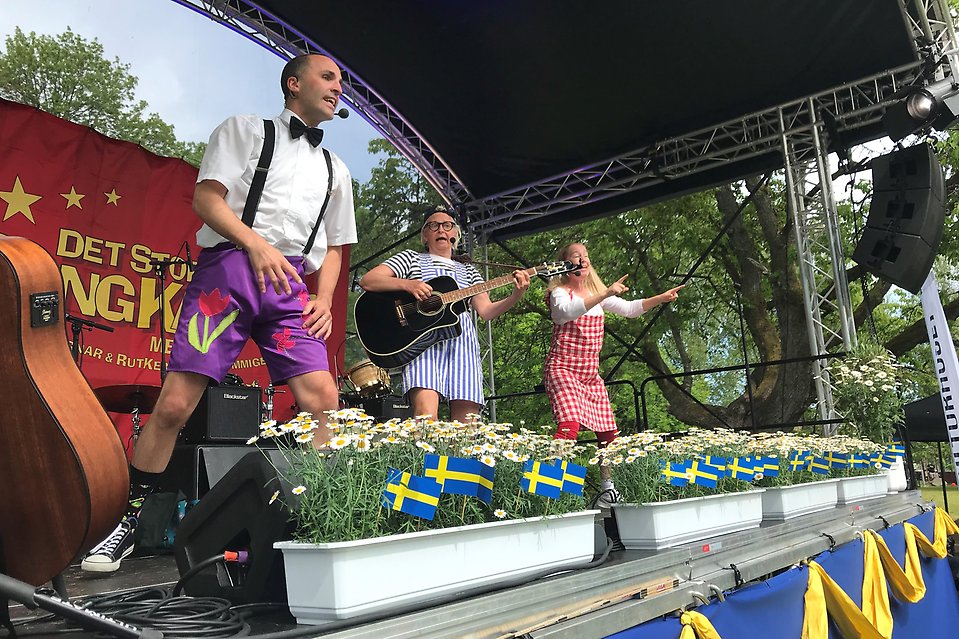 Tre personer står på scenen och uppträder. En man i vit skjorta och svart fluga sjunger, en kvinna i blårandig klänning spelar gitarr och en kvinna i rödrutig klänning dansar.