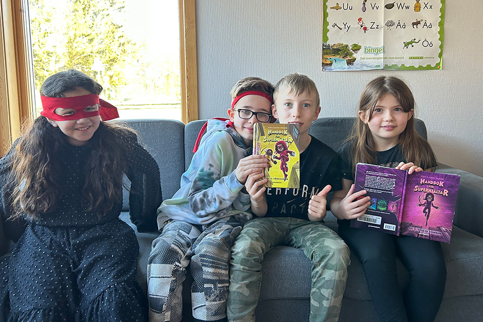 Fyra barn sitter i en soffa med två böcker framför sig. De ler in i kameran och en tjej har en röd mask på sig.