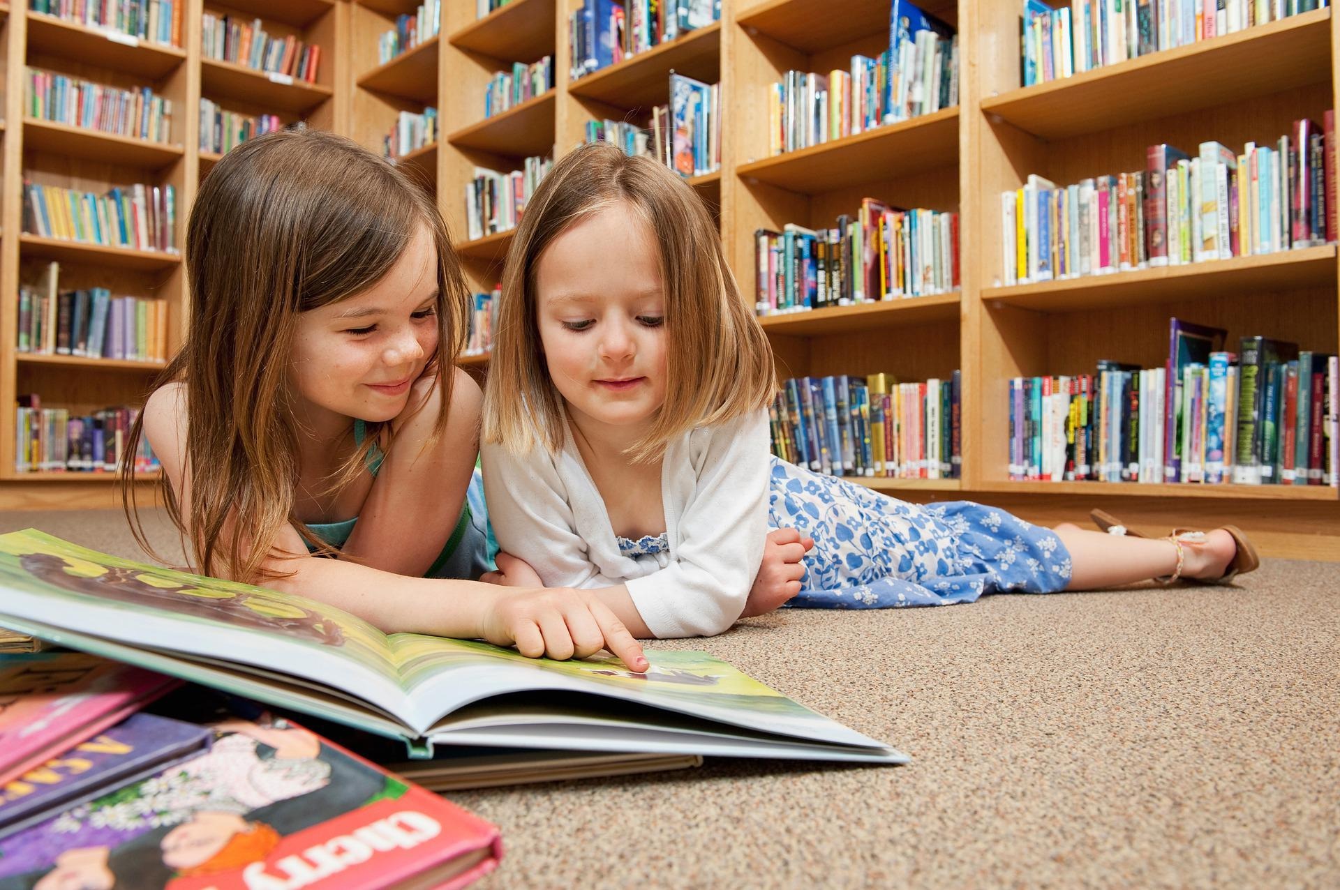 Två barn ligger på mage på golvet inne i biblioteket och läser en bok tillsammans