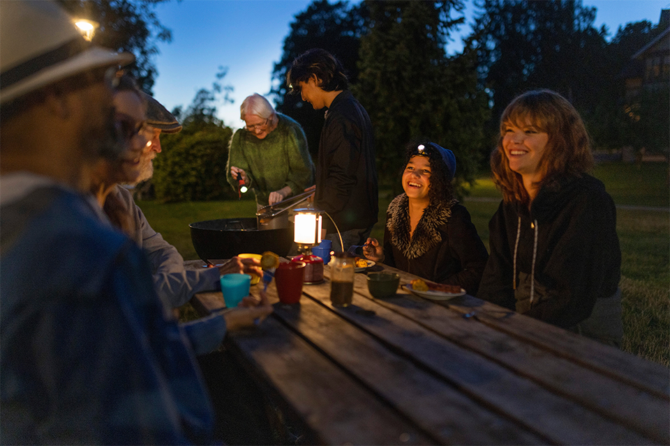 Människor vid ett bord utomhus. I bakgrunden en grill. Det mörknar.