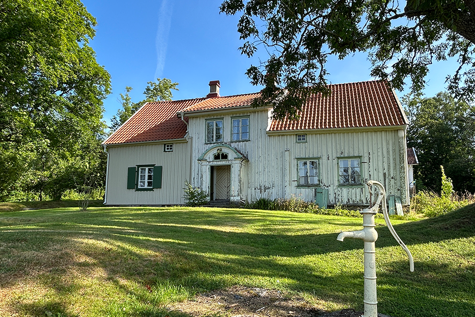 Vitt äldre hus. I förgrunden en gammal vattenpump. Huset omgärdat av träd.