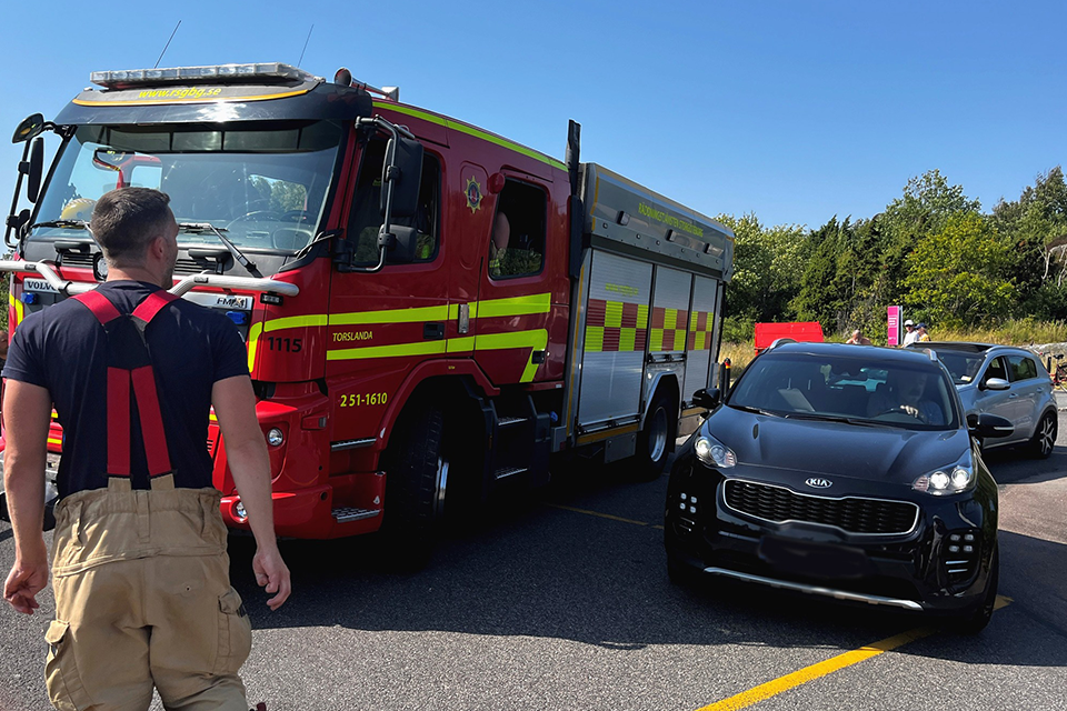 Brandbil bredvid parkerade bilar en solig dag. En brandman i förgrunden.
