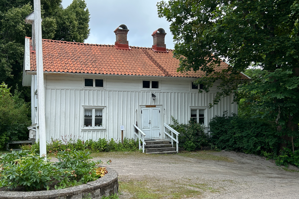 Äldre vitt hus. I förgrunden en flaggstång, bakom huset träd.