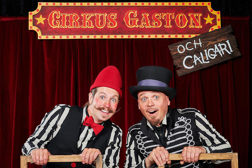 Två män utklädda i cirkuskläder sitter på varsin stol bak och fram. På en skylt står Cirkus Gaston och Calgigari