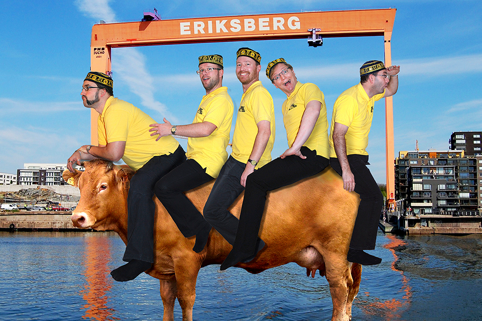 Fem glada män i gult rider på en ko. I bakgrunden Eriksbergskranen.