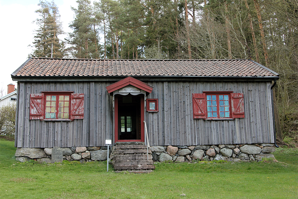 Äldre grå trästuga med rött runt fönstren. I bakgrunden skog.