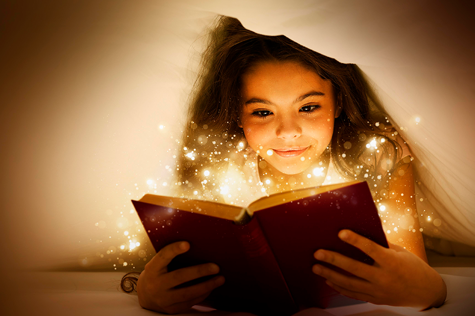 Flicka ligger under täcket och läser en bok där ljus strömmar ut över hennes ansikte från boken.