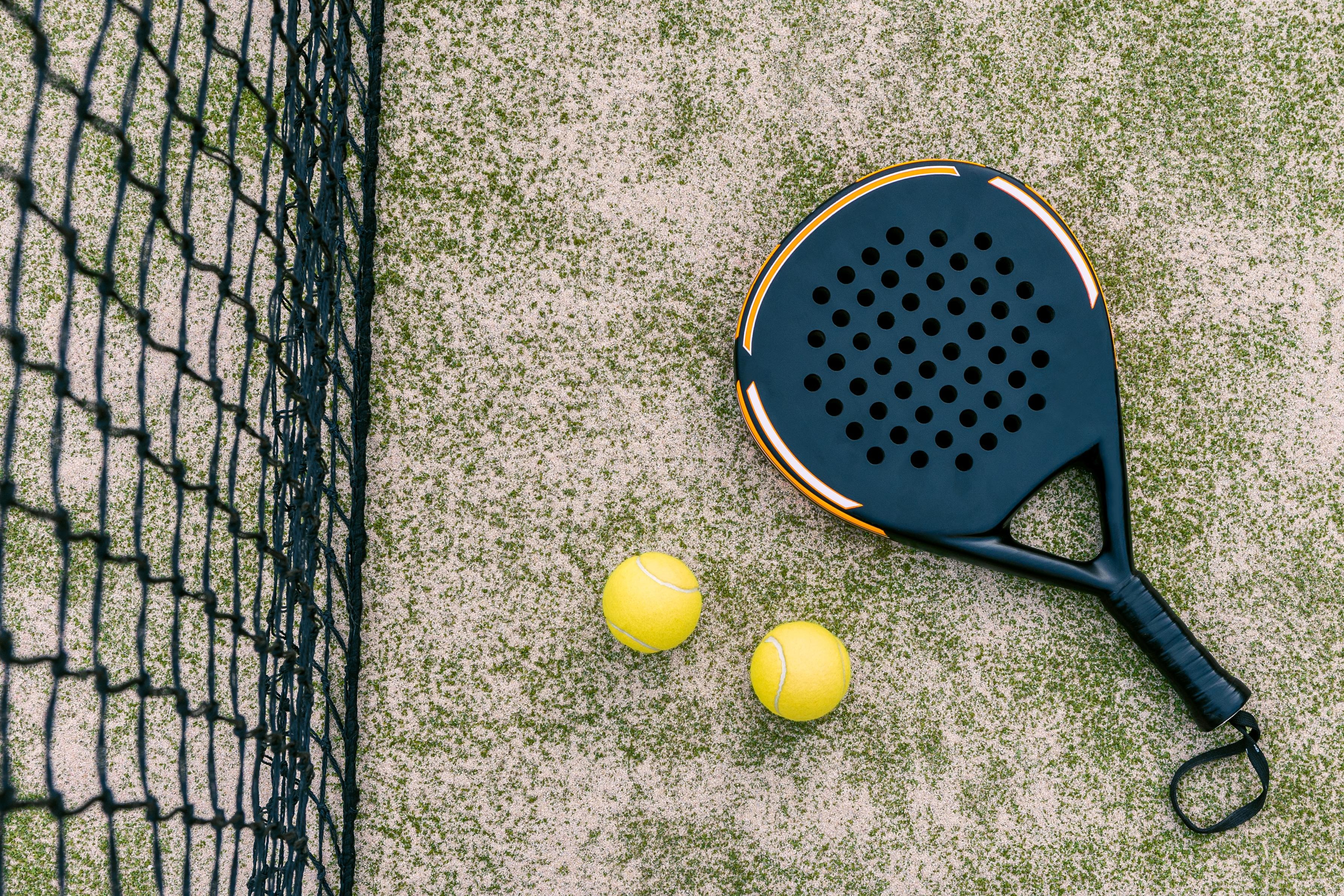 Bilden är tagen ovanifrån. Ett paddelrack och två tennisbollar ligger på en tennisbana.