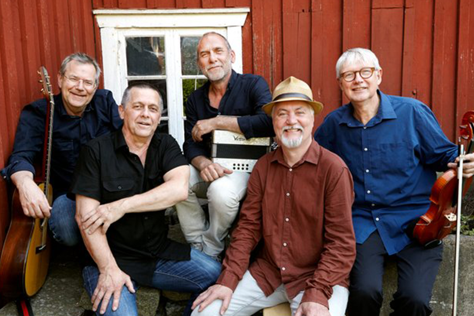 Fem herrar med instrument står mot en rödmålad, äldre trävägg. De ser glada ut.