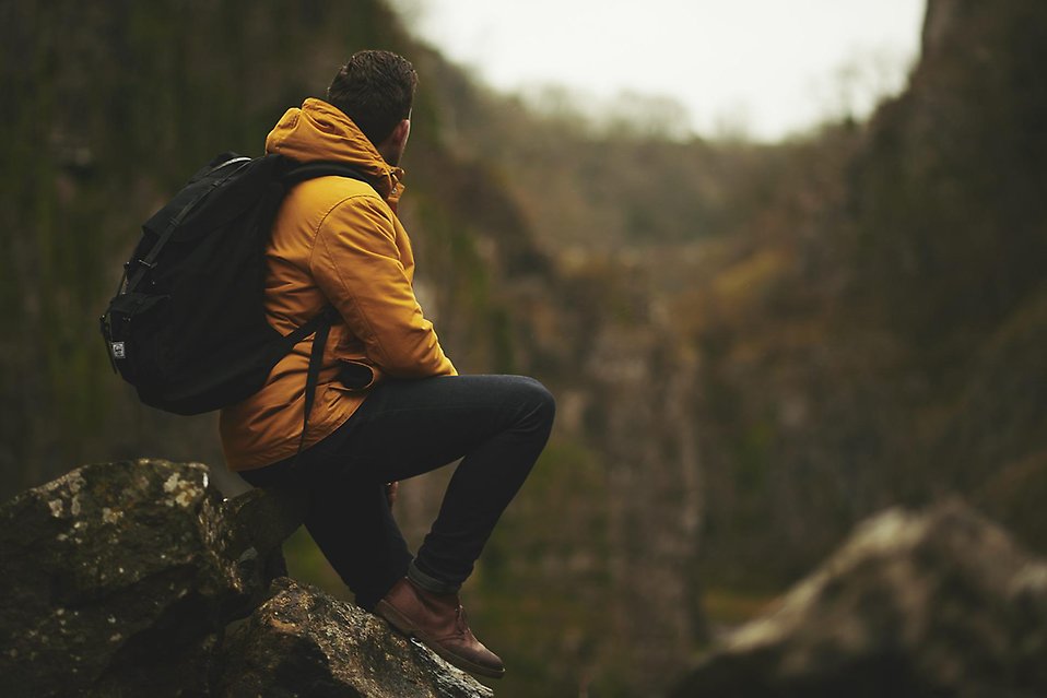 En man i gul jacka och svart ryggsäck sitter med ryggen mot kameran och en vandringstig syns i bakgrunden