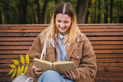 Ung flicka sitter på en parkbänk och läser och skrattar.