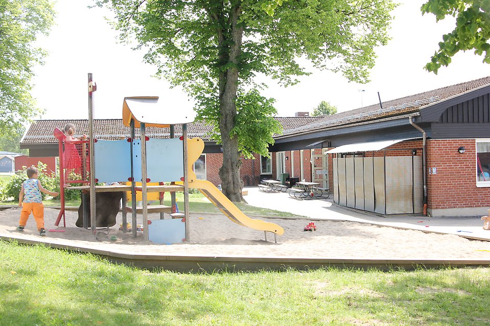 Lekplats med klättertorn och rutchelkana där två barn leker. I bakgrunden syns ett rött enplanshus