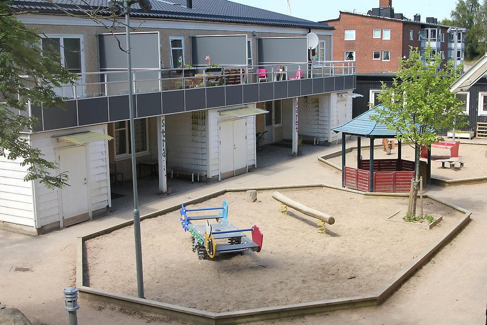 En stor lekplats med sandlåda och lusthus och bakom står ett vitt tvåvåningshus med balkonger på andra våningen