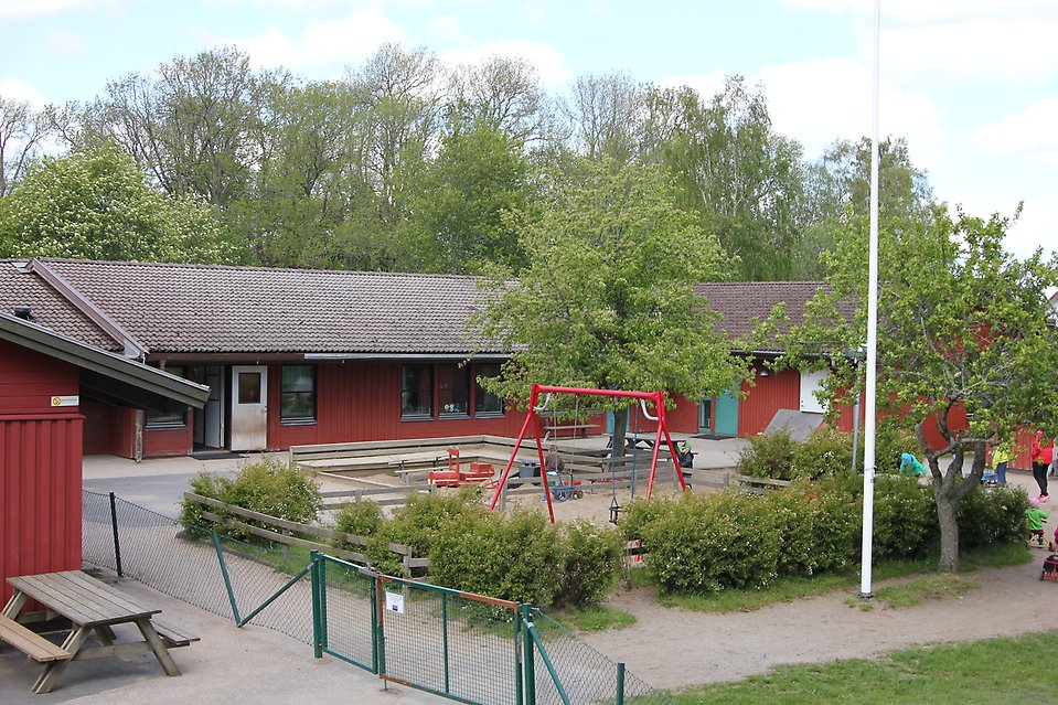 Inhägnad lekplats med röd gungställning och sandlåda. Bakom står ett U format rött hus.