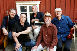 Fem herrar med instrument står mot en rödmålad, äldre trävägg. De ser glada ut.