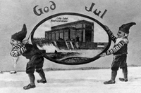 Svartvit bild, två tomtar bär en rund tavla med Lilla Edets kraftstation. Äldre motiv, grafik "God Jul".