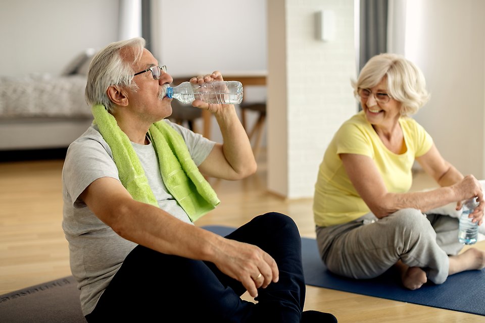 Två äldre personer sitter på varsin träningsmatta och dricker vatten