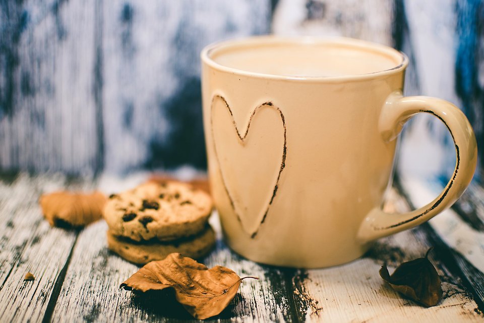 En vit kaffekopp med ett hjärta på. Framför koppen ligger två kakor och några höstlöv
